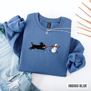 Puede incluir: Sudadera azul &iacute;ndigo con un perro de dibujos animados persiguiendo a un mu&ntilde;eco de nieve. El perro es negro y el mu&ntilde;eco de nieve tiene una bufanda roja. La sudadera tiene cuello redondo y un collar plateado. El texto "INDIGO BLUE" est&aacute; en la parte inferior.