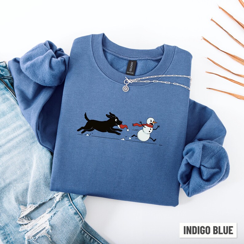 Puede incluir: Sudadera azul &iacute;ndigo con un perro de dibujos animados persiguiendo a un mu&ntilde;eco de nieve. El perro es negro y el mu&ntilde;eco de nieve tiene una bufanda roja. La sudadera tiene cuello redondo y un collar plateado. El texto "INDIGO BLUE" est&aacute; en la parte inferior.