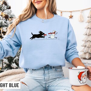 Puede incluir: Sudadera azul claro con un perro negro persiguiendo a un mu&ntilde;eco de nieve con una bufanda roja. El texto "LIGHT BLUE" est&aacute; en la parte inferior. El dise&ntilde;o es un gr&aacute;fico divertido con tem&aacute;tica invernal.