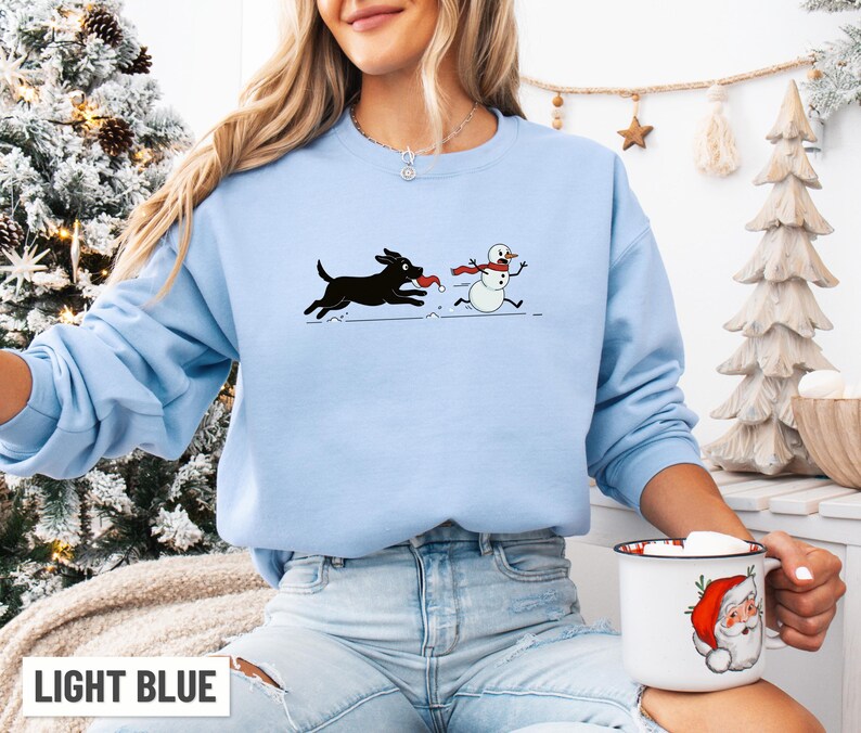 Puede incluir: Sudadera azul claro con un perro negro persiguiendo a un mu&ntilde;eco de nieve con una bufanda roja. El texto "LIGHT BLUE" est&aacute; en la parte inferior. El dise&ntilde;o es un gr&aacute;fico divertido con tem&aacute;tica invernal.