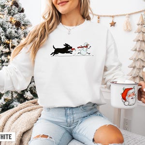 Puede incluir: Sudadera blanca con un perro negro persiguiendo a un mu&ntilde;eco de nieve, ambos con bufandas rojas. Una taza blanca con un dise&ntilde;o de Pap&aacute; Noel se sostiene en la mano. La palabra "WHITE" est&aacute; impresa en la parte inferior.