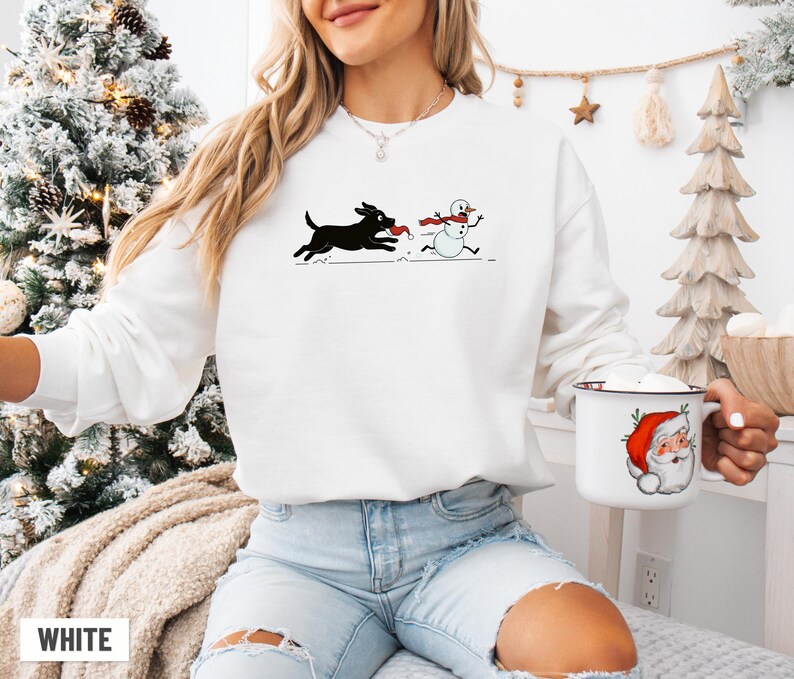 Puede incluir: Sudadera blanca con un perro negro persiguiendo a un mu&ntilde;eco de nieve, ambos con bufandas rojas. Una taza blanca con un dise&ntilde;o de Pap&aacute; Noel se sostiene en la mano. La palabra "WHITE" est&aacute; impresa en la parte inferior.