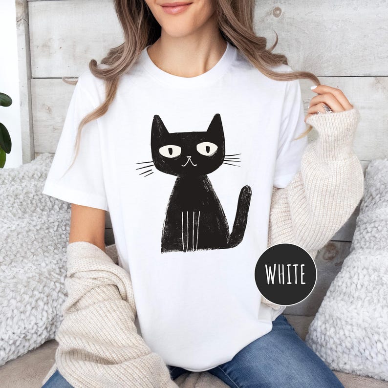 Pu&ograve; includere: Maglietta bianca con un'illustrazione di un gatto nero. Il gatto &egrave; seduto con la coda avvolta intorno alle zampe. Il gatto ha un design semplice e cartonato con occhi grandi e una piccola bocca.
