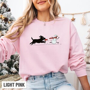 Puede incluir: Sudadera rosa claro con un dibujo animado de un perro negro persiguiendo a un mu&ntilde;eco de nieve. El perro lleva un gorro de Pap&aacute; Noel y el mu&ntilde;eco de nieve tiene una bufanda roja. El texto "LIGHT PINK" est&aacute; en la parte inferior. Un dise&ntilde;o festivo.