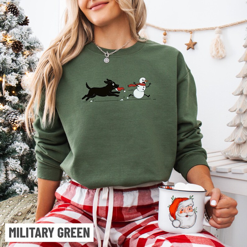 Puede incluir: Sudadera verde militar con un gr&aacute;fico de un perro negro persiguiendo a un mu&ntilde;eco de nieve. El perro lleva un juguete rojo en la boca. El mu&ntilde;eco de nieve lleva una bufanda roja. El texto "MILITARY GREEN" est&aacute; en la parte inferior. Tambi&eacute;n se ve una taza blanca con un dise&ntilde;o de Pap&aacute; Noel.