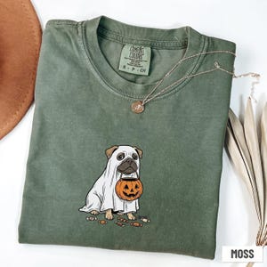 May include: A moss green short-sleeve t-shirt featuring a graphic of a pug wearing a white ghost costume and holding an orange jack-o'-lantern candy bucket. Small candy pieces are scattered on the ground around the dog. The shirt has a Comfort Colors brand label at the inner neckline.