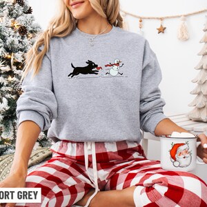 Puede incluir: Sudadera gris deportiva con un perro negro persiguiendo a un mu&ntilde;eco de nieve con una bufanda roja. La sudadera se combina con pantalones de pijama a cuadros rojos y blancos. Se sostiene una taza blanca con un dise&ntilde;o de Pap&aacute; Noel.