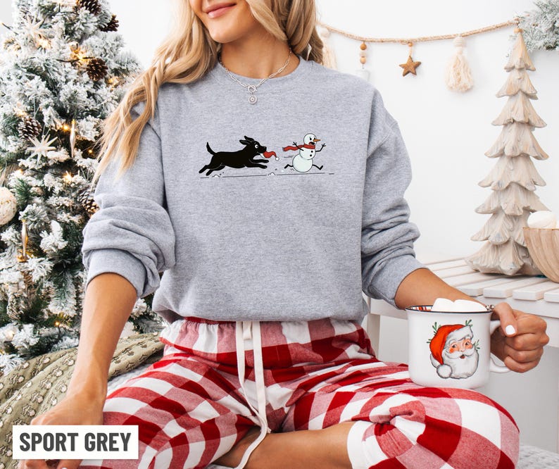 Puede incluir: Sudadera gris deportiva con un perro negro persiguiendo a un mu&ntilde;eco de nieve con una bufanda roja. La sudadera se combina con pantalones de pijama a cuadros rojos y blancos. Se sostiene una taza blanca con un dise&ntilde;o de Pap&aacute; Noel.