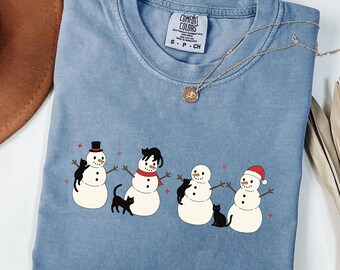 Puede incluir: Camiseta Comfort Colors azul vaquero con cuatro muñecos de nieve y gatos negros. Cada muñeco de nieve lleva un sombrero o bufanda diferente. El texto "BLUE JEAN" está en la parte inferior.