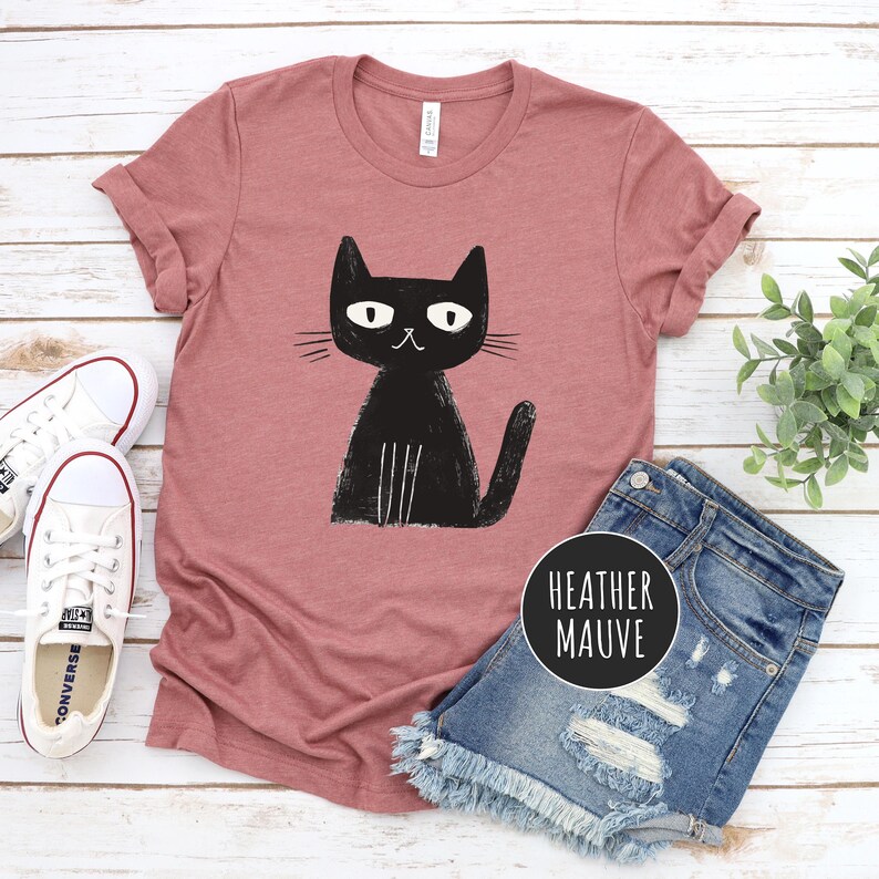 Pu&ograve; includere: Una t-shirt color malva sfumato con un'immagine di un gatto nero. Il gatto &egrave; seduto con la coda arrotolata intorno alle zampe. La t-shirt &egrave; su una superficie di legno bianco.