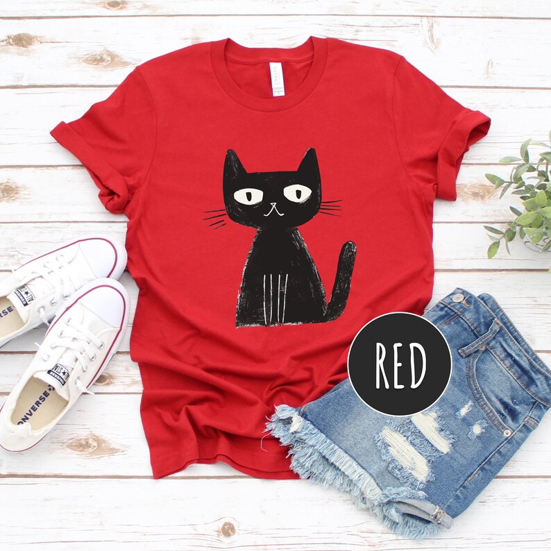 Pu&ograve; includere: Maglietta rossa con un gatto nero stampato. Il gatto &egrave; seduto e guarda a sinistra. La parola "ROSSO" &egrave; scritta in bianco su un cerchio nero.