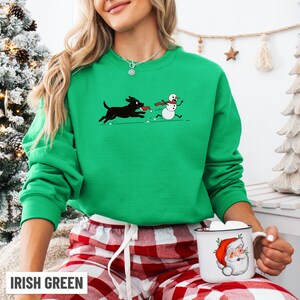 Puede incluir: Sudadera verde irland&eacute;s con un dibujo animado de un perro negro persiguiendo a un mu&ntilde;eco de nieve. El perro tiene un juguete rojo, y el mu&ntilde;eco de nieve lleva una bufanda roja. El texto "IRISH GREEN" est&aacute; en la parte inferior. Cuello redondo y mangas largas.