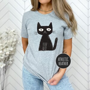 Pu&ograve; includere: Una maglietta grigia con un'immagine di un gatto nero. Il gatto ha un design semplice e cartonato con occhi grandi e una piccola bocca. Il testo "Athletic Heather" &egrave; visibile nell'angolo in basso a destra della maglietta.