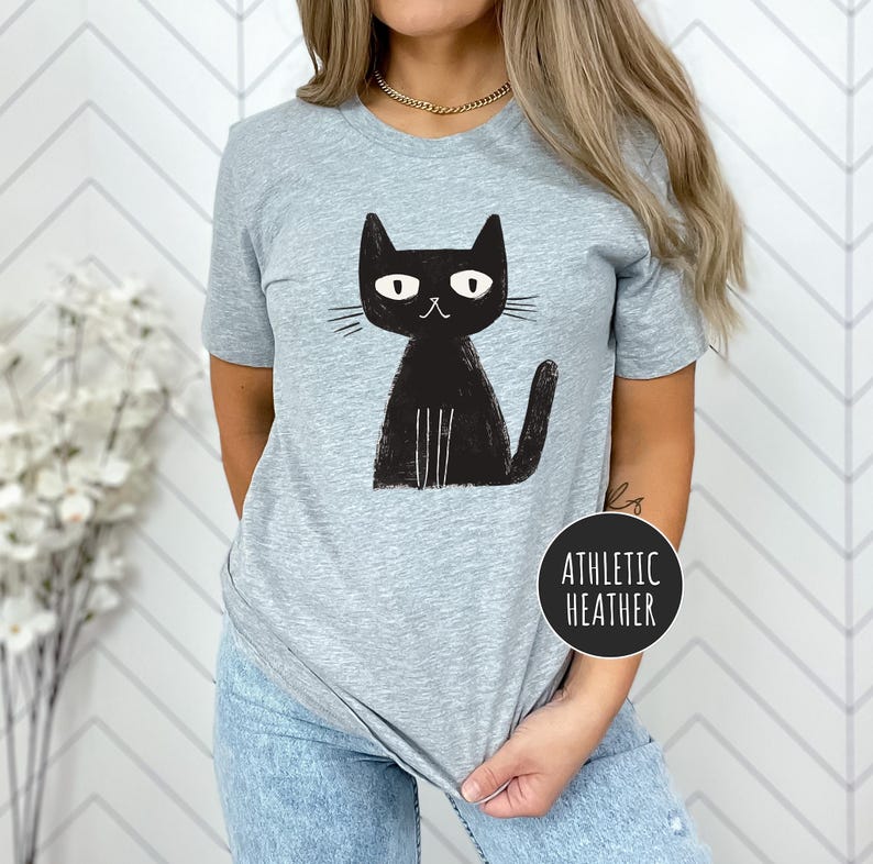 Pu&ograve; includere: Una maglietta grigia con un'immagine di un gatto nero. Il gatto ha un design semplice e cartonato con occhi grandi e una piccola bocca. Il testo "Athletic Heather" &egrave; visibile nell'angolo in basso a destra della maglietta.