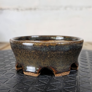 Peut inclure: Petit pot à bonsaï en céramique fait à la main, avec une glaçure sombre et mouchetée. Le pot en forme de bol a une large ouverture et repose sur quatre pieds. La couleur est un mélange de marron, de noir et de reflets bleus, avec une finition brillante.