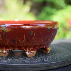 May include: A round, low-profile ceramic bonsai pot with a glossy, deep red glaze featuring vertical, dripping brown and gold accents. The vessel is supported by four short, unglazed, light brown feet that elevate the base from the surface.