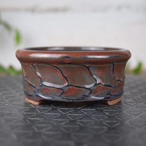 以下が含まれることがあります： 茶色、黒、青の色合いの、質感のあるひび割れ模様の小さな丸いセラミック盆栽鉢。縁は濃い茶色で、鉢は小さな足の上にあります。この鉢は小さな植物用に設計されています。