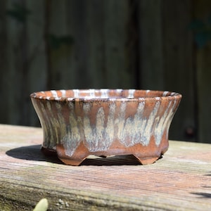 May include: A round ceramic bonsai pot with a flared rim and four small feet. The exterior features a glossy glaze with a blend of earthy brown tones and vertical streaks of cream and light gray dripping down from the top edge to the base.