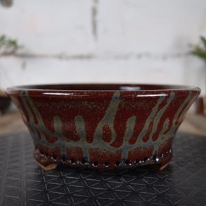 Peut inclure: Un pot à bonsaï en céramique émaillée de couleur brun rougeâtre. Le pot présente une forme évasée et est orné d'un motif abstrait et fluide dans des tons gris et bleus. Le pot mesure environ 20 cm de diamètre.