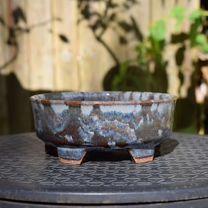 May include: A round ceramic bonsai pot featuring a reactive glaze with blended tones of deep brown, slate blue, and white. The vessel has a wide, open top, a thick rim, and is supported by four short, sturdy feet at the base, showcasing a textured finish.