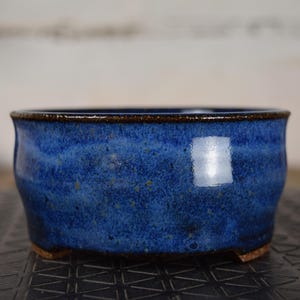 Puede incluir: Una maceta de bonsái de cerámica azul hecha a mano con un esmalte moteado. El recipiente redondo y de perfil bajo tiene un borde marrón oscuro y pequeños pies. La maceta está diseñada para plantar árboles y arbustos pequeños.