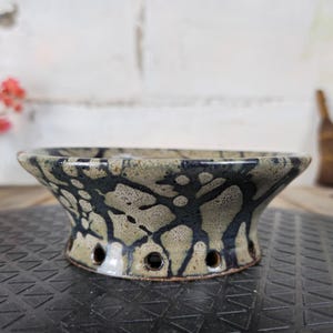 以下が含まれることがあります： 青とベージュの色合いの、フレアベースと斑点のある有機的な模様が特徴のセラミックボウル。ボウルの底には小さな穴がいくつかあります。このボウルは、装飾または機能的な目的で使用される可能性があります。