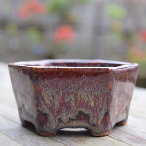 May include: A small, square bonsai pot with a rich, glossy, reddish-brown glaze. The ceramic pot has a slightly textured surface and sits on three small feet. Ideal for miniature plants, it measures approximately 5 inches wide.