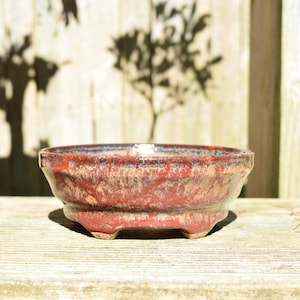 Może przedstawiać: Okrągła ceramiczna doniczka do bonsai z błyszczącym szkliwem w odcieniach głębokiej czerwieni, ziemistego brązu i subtelnej czerni. Naczynie ma szeroki otwór, lekko zwężające się ścianki i jest wsparte na czterech małych, zintegrowanych nóżkach.