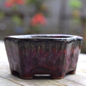 Könnte beinhalten: Ein quadratischer Bonsai-Topf mit dunkler, gesprenkelter Glasur in Burgunder-, Braun- und Schwarztönen. Der Keramiktopf hat eine leicht strukturierte Oberfläche und steht auf vier kleinen Füßen. Der Rand ist heller.