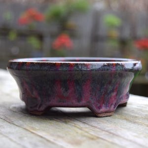 May include: A small, ceramic bonsai pot with a dark, glossy finish. The pot features a deep burgundy and black color scheme with a textured appearance. It has a low, wide shape with three small feet.
