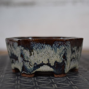 Peut inclure: Un pot à bonsaï carré avec un bord marron foncé et un extérieur vernissé texturé. Le vernis présente un mélange de teintes blanches, bleues et marron, créant un aspect artistique unique. Le pot repose sur de petits pieds bruns.