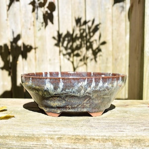 May include: A shallow, round ceramic bonsai pot with a wide rim and three small feet. The exterior features a reactive glaze with a deep brown base, transitioning into streaks of white and light blue that drip down the sides toward the base.