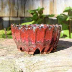 May include: A round ceramic bowl with a deep red, glossy glaze and a textured, fluted exterior. The rim features a scalloped edge, and the base has a series of arched cutouts, revealing the natural, unglazed brown clay underneath.