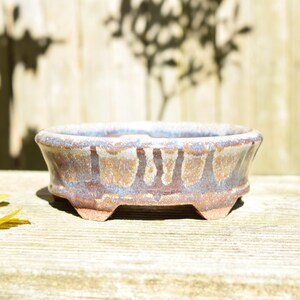 May include: A shallow, oval-shaped ceramic bonsai pot featuring a reactive glaze with blended tones of earthy brown, muted blue, and cream. The pot has a wide, flared rim, a smooth interior, and is supported by four small, sturdy, integrated feet.
