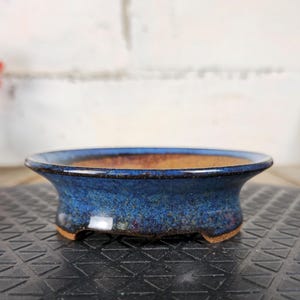 Peut inclure: Un pot à bonsaï rond en céramique bleue avec un glaçage moucheté. Le pot a un design large et peu profond avec un intérieur marron et de petits pieds. Le bord est bleu foncé et le pot repose sur une surface texturée.