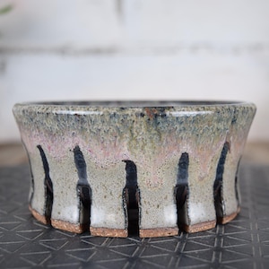 Peut inclure: Un bol en céramique fait à la main avec un design unique. Le bol présente un glaçage gris et rose avec des rayures noires verticales. Le bord est légèrement évasé et la base a une finition naturelle non émaillée. Idéal pour les plantes ou la décoration.