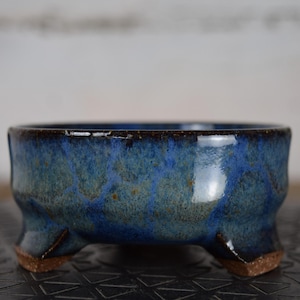 Peut inclure: Un bol en céramique bleu fait à la main avec une finition brillante. Le bol a un profil bas, une forme arrondie et trois petits pieds. L'extérieur présente un motif moucheté de bleus et de verts, avec un bord et des fonds de pieds brun foncé.