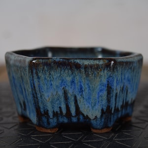 Peut inclure: Un pot à bonsaï carré en céramique avec un glaçage bleu et marron. Le pot a une surface texturée et repose sur trois petits pieds. Le glaçage a un effet dégoulinant, créant un attrait visuel unique. Idéal pour les petites plantes.