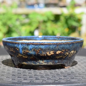 May include: A shallow, round ceramic bonsai pot featuring a deep blue and black reactive glaze with visible brown earthy undertones. The vessel has a thick, slightly uneven rim and is supported by three short, sturdy feet at the base.