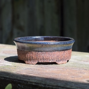 May include: A round ceramic bonsai pot featuring a textured, reddish-brown clay base with vertical grooves and a smooth, glossy dark blue and black glazed rim. The vessel has a wide, open top and sits on small, integrated feet at the bottom.