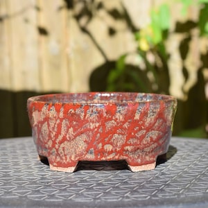 May include: A round ceramic bonsai pot featuring a textured glaze with a mottled pattern of deep red, brown, and tan tones. The vessel has a wide, open top, a slightly tapered body, and four small, sturdy feet at the base for elevation.