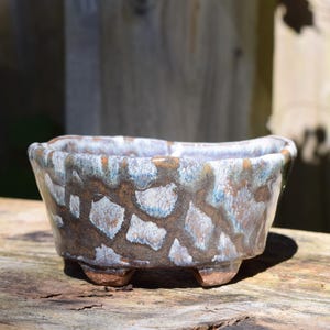 May include: A small, oval-shaped ceramic bonsai pot featuring a textured, multi-colored glaze with a pattern of brown, white, and light blue patches. The vessel has a slightly uneven, wavy rim and is supported by four short, sturdy feet at the base.