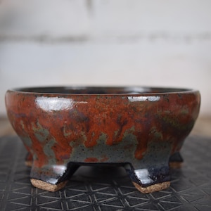 Peut inclure: Un pot à bonsaï en céramique aux couleurs riches et terreuses. Le bol présente un mélange de teintes rouges, brunes et bleu-gris, avec une finition brillante. Il a un profil bas et est soutenu par trois pieds robustes.