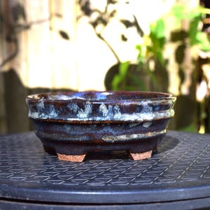 May include: A round ceramic bonsai pot with a dark brown and deep blue glaze featuring a mottled, light-colored rim. The pot has a wide, shallow basin, a slightly flared top edge, and stands on four small, unglazed, terracotta-colored feet.