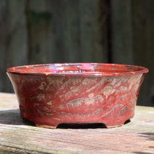 May include: A round, shallow ceramic bonsai pot with a glossy, mottled red and brown glaze. The exterior features a textured, streaked finish with darker brown accents. The pot has a wide, open top and sits on four small, integrated feet.