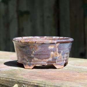 Może przedstawiać: Okrągła ceramiczna doniczka do bonsai z nakrapianym brązowo-niebieskim szkliwem. Naczynie posiada lekko rozszerzany brzeg, teksturowaną powierzchnię zewnętrzną i stoi na czterech małych, zintegrowanych nóżkach u podstawy, co nadaje mu rustykalny wygląd.