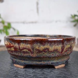 May include: A ceramic bonsai pot with a wide, shallow design. The pot features a rich, glazed finish with a blend of brown, red, and yellow hues. It has a slightly textured surface and is supported by three small feet. The pot is designed for bonsai trees.