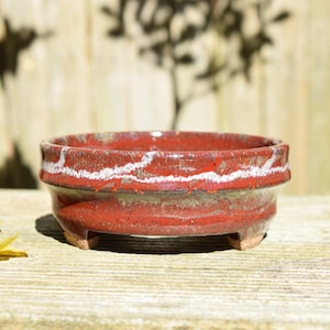 May include: A round ceramic bonsai pot with a glossy red glaze and a textured white drip pattern along the upper rim. The pot features a slightly flared edge, a deep basin, and stands on three short, sturdy feet, showcasing a rustic finish.