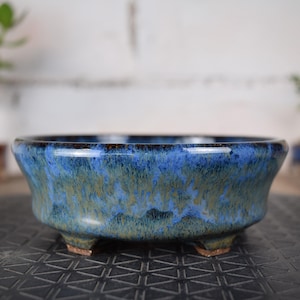 Peut inclure: Un pot à bonsaï en céramique émaillée avec une finition mouchetée bleue et verte. Le pot rond a trois petits pieds et un bord foncé. Le pot est conçu pour contenir de petites plantes.