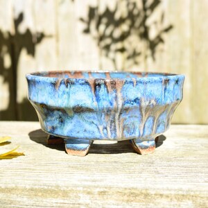 May include: A round ceramic bonsai pot featuring a vibrant blue glaze with brown drip accents. The bowl has a faceted exterior surface and is supported by four short, sturdy feet. The rim is slightly uneven, showcasing a blend of blue and brown tones.